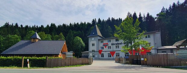 die Strobel-Mühle von der Talstraße aus gesehen: Einfahrt, Kapelle, ein grüner Baum und das Haus in Grautönen und dem roten CVJM-Dreieck